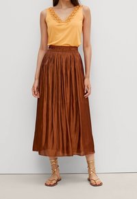Top giallo senza maniche con pizzo, abbinato a una lunga gonna plissettata marrone. Il modello indossa sandali con cinturino, mostrando un look estivo rilassato.