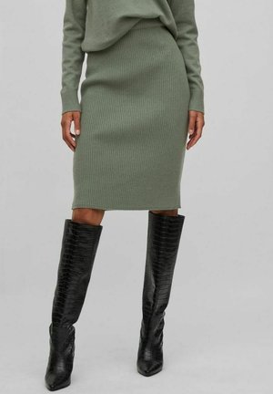 Femme portant une jupe côtelée olive longueur genou et des bottes en cuir noires texturées montant jusqu'aux genoux, debout devant un fond gris uni.