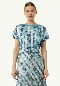 Niebieski top w stylu tie-dye o luźnym kroju i krótkich rękawach, z okrągłym dekoltem. Dobrany do spódnicy w tym samym stylu tie-dye. Gładka tekstura materiału.