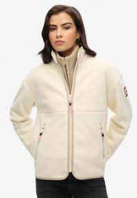 Witte fleecejack met een ritssluiting aan de voorkant, twee zijzakken en een hoge kraag. Heeft roze accenten op de ritsen. Glad textuur.