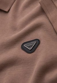 Tejido de camiseta polo marrón con una textura suave. Presenta un parche triangular negro con "OMBRA EXIMIO" grabado en plata.