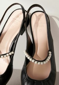 Mustat nahkaiset slingback-kengät, joissa on pyöristetty kärki, helmikoristeinen hihna ja matala tukikorko. Pehmeä vaaleanpunainen pohjallinen näkyy.