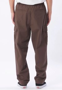 Man med bruna cargobyxor med elastisk midja, bakfickor med bál och sidofickor med flik, som är kombinerade med svartvita sneakers.