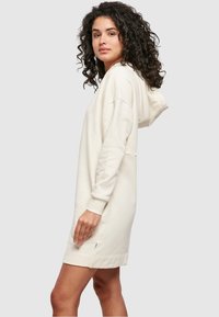 Krēmkrāsas kapuces svārku kleita ar gariem piedurknēm, sānu kabatām un mīkstu faktūru. Tam ir brīvs piegriezums un ribots aproces.