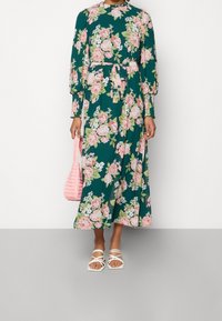 Vestido maxi floral en verde con rosas rosas, mangas largas, cintura ajustada y un cinturón a juego. Combinado con un bolso de mimbre rosa y sandalias blancas.