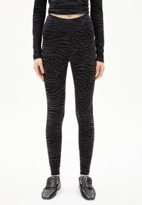 Vrouw in zwart en donkergrijs met zebra-strepen leggings, bijpassende lange mouwen top en zwarte geprikte instappers, staand op een witte achtergrond.