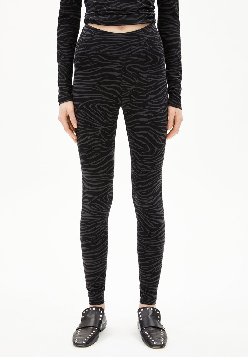 Vrouw in zwart en donkergrijs met zebra-strepen leggings, bijpassende lange mouwen top en zwarte geprikte instappers, staand op een witte achtergrond.