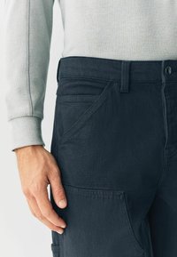 Dark blue cargo pants with a textured fabric, featuring multiple pockets, a button closure, and a straight cut, paired with a light grey top.