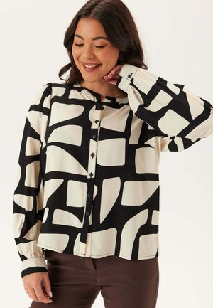 Kvinna som bär en långärmad blus med knappar och ett svart och krämfärgat abstrakt geometriskt mönster, matchat med mörkbruna byxor, ler med handen nära nacken.