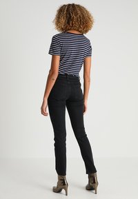 Jean taille haute noirs associés à un t-shirt rayé bleu marine et blanc. Le modèle porte des bottines à talons métalliques. Cheveux bouclés visibles.