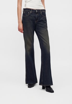 Person iført mørke vide jeans med let falmning og sorte hælede sko, står mod en ensfarvet lys baggrund.
