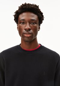 Felpa nera con scollo rotondo, caratterizzata da un logo discreto sul petto. Indossata sopra una maglietta rossa, il tessuto appare liscio e morbido.