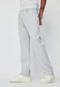Pantalon gris clair avec un design à jambes droites, doté de plis à l'avant, associé à des baskets blanches. Tissu lisse et coupe ajustée.