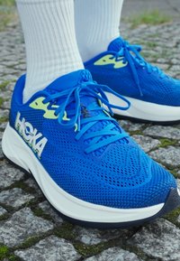Blauwe sportieve schoenen met een ademend bovenwerk van mesh, contrasterende gele accenten, platte veters en een gewatteerde witte tussenzool met een gestructureerde buitenzool.