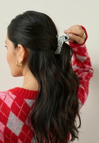 Kvinna med långt mörkt hår som bär guldörhängen och en röd och rosa mönstrad tröja, håller en svart och vit fläckig hårklämma.