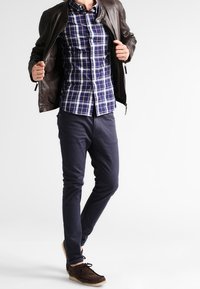 Brązowa skórzana kurtka na niebiesko-białej koszuli w kratę, do tego granatowe spodnie i brązowe zamszowe buty. Prosty, casualowy styl.