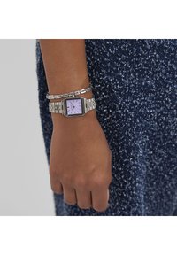 Silverfärgad metallklocka med fyrkantig urtavla och lavendelfärgad dial, tillsammans med ett länkarmat silverarmband, buren på en hand mot en mörk texturerad tyg.