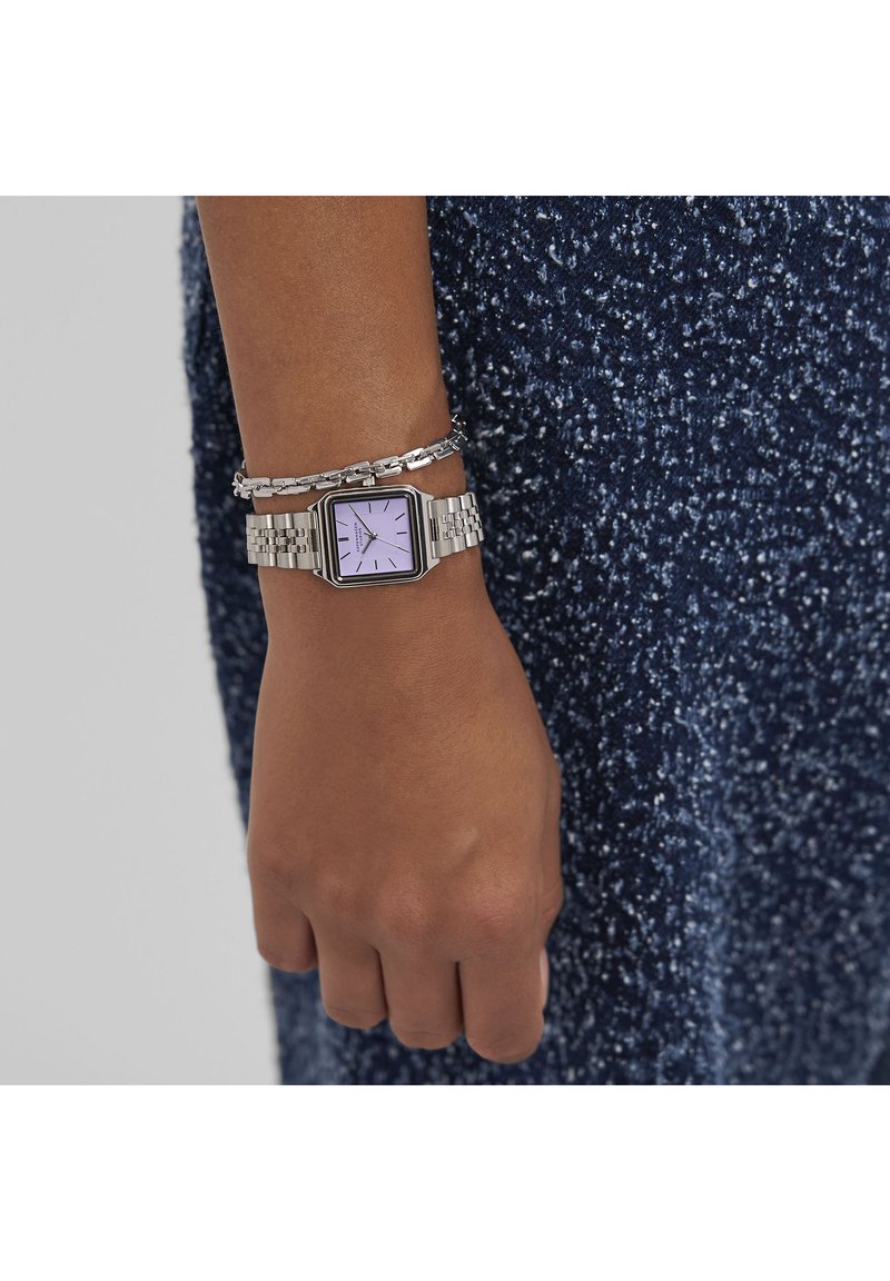 Silverfärgad metallklocka med fyrkantig urtavla och lavendelfärgad dial, tillsammans med ett länkarmat silverarmband, buren på en hand mot en mörk texturerad tyg.