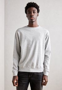 Sudadera gris claro con cuello redondo, mangas largas y puños acanalados. El tejido parece suave con una textura sutil. Jeans negros abajo.