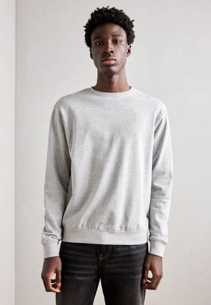 Sweatshirt gris clair avec un col rond, des manches longues et des poignets côtelés. Le tissu semble doux avec une texture subtile. Jeans noirs en dessous.