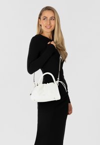 Bolso acolchado blanco con forma estructurada, asas redondas y correa de plata ajustable. Presenta un sutil branding en la parte frontal.