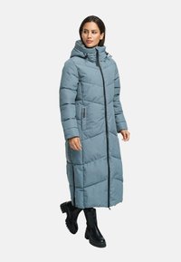 Woman wearing a long blue padded winter coat with hood, black zipper, side pockets, and black chunky boots, standing and looking sideways.