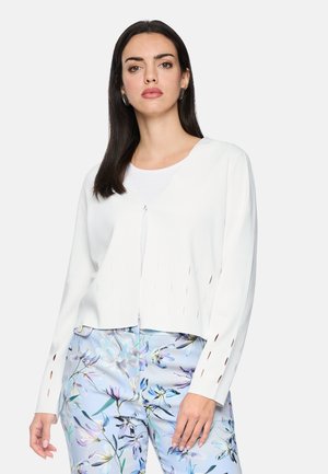 Frau mit langen dunklen Haaren, die ein weißes langärmliges Oberteil und hellblaue Hosen mit Blumenmuster trägt, steht vor einem einfarbigen Hintergrund.