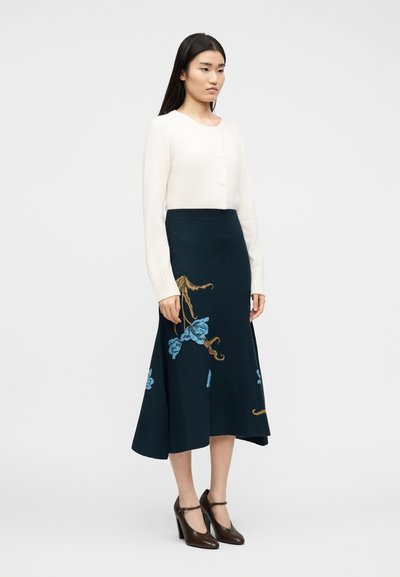 Γυναίκα που φοράει λευκή μακρυμάνικη ζακέτα, μπλε ναυτικό μίντι φούστα με μπλε και καφέ ανθιστή κεντήματα, και σκούρα καφέ παπούτσια με τακούνια.