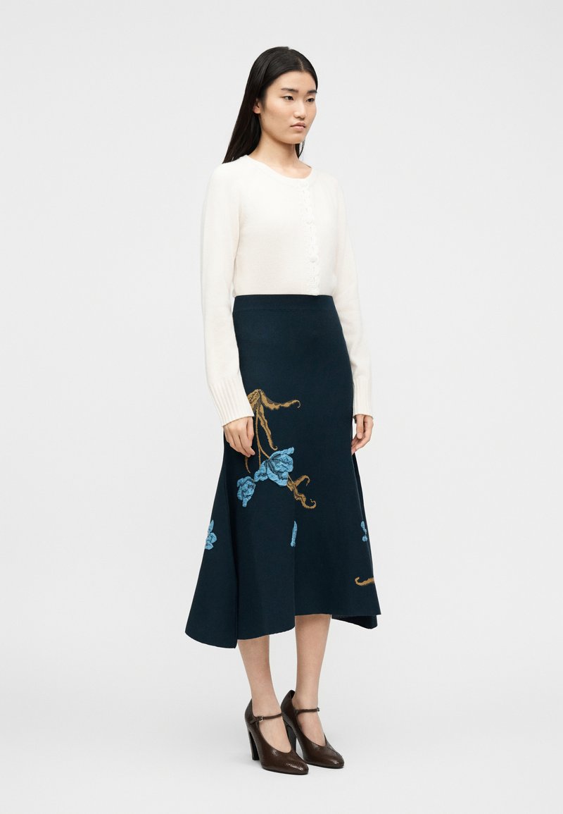 Vrouw draagt een witte cardigan met lange mouwen, een marineblauwe midirok met blauw en bruin bloemenborduursel, en donkerbruine schoenen met hak.