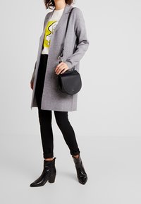 Šedý dlouhý kardigan přes bílou košili s žlutou grafikou, v kombinaci s černými slim kalhotami a černými kotníkovými botami. Zobrazená je černá crossbody taška.