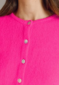 Moteris dėvi ryškiai rožinį pūstą megztinį su keturiais matomais perlamutro sagomis, atidengiančią kaklą ir viršutinę krūtinės dalį.