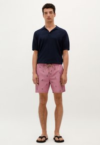 Polo cu mânecă scurtă în nuanță de marină, pantaloni de baie roz cu model de cranii negre, talie cu șnur, șlapi negri, modelul stând în fața unui fundal simplu.