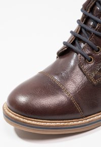 Hnědá kožená bota s černými tkaničkami, světle hnědou šitým podrážkou a podrobným šitím na špičce a po stranách, umístěná na bílém pozadí.