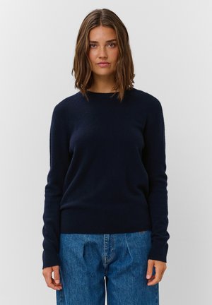 Giovane donna con capelli castani fino alle spalle che indossa un maglione blu scuro e jeans blu, in piedi davanti a uno sfondo chiaro e uniforme.