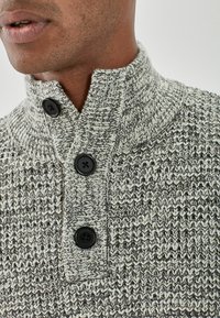 Gebreide grijze en crèmekleurige trui met een hoge kraag, voorzien van drie zwarte knopen en een gestructureerd patroon in de stof.