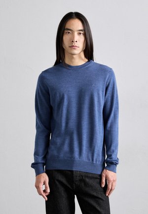 Blauer Strickpullover mit rundem Halsausschnitt sowie gerippten Bündchen und Saum. Getragen mit dunklen Jeans. Zartes Logo auf der Brust. Glatte Textur.