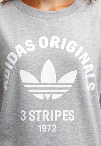 Grå sweatshirt med vit Adidas Originals-logotyp, trefalksymbol och texten "3 Stripes 1972" på framsidan.