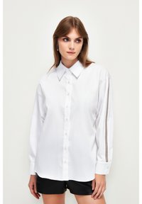Camicia bianca a maniche lunghe con colletto a punta, chiusura frontale a bottoni e due strisce verticali grigie sulle maniche. Tessuto liscio, vestibilità relax.