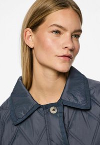 Jūrų mėlynos spalvos siuvinėtas švarkas su dideliu apykakle ir mygtukų uždarymu. Lygus, blizgus paviršius su įstrižais siuvinėjimo raštais.