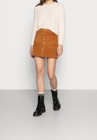 Cord-Minirock in goldbraun mit Knopfleiste, kombiniert mit einem cremefarbenen, strukturierten Strickpullover und schwarzen Schnürstiefeln im Combat-Stil.