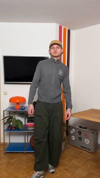 Νεαρός άνδρας με γκρι πουλόβερ με φερμουάρ, ελιά παντελόνι φαρδύ, μπεζ καπέλο, στέκεται σε δωμάτιο με ριγέ τοίχο, πορτοκαλί λάμπα και βαλίτσα με αυτοκόλλητα.