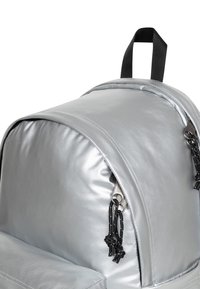 Szary plecak z zaokrąglonym wierzchem, wykonany z błyszczącego materiału. Posiada czarne suwaki oraz akcenty sznurków na suwakach.