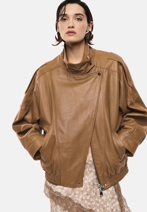 Donna con capelli scuri che indossa grandi orecchini a forma di cuore, una giacca di pelle marrone e un vestito floreale rifinito con pizzo, mani nelle tasche della giacca.