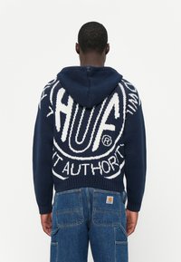Marineblå strikket hettegenser med hvite grafikker som sier "HUF" og "AUTHORITY" på ryggen; ribbestrikket kant og mansjetter, med snor i hetten.