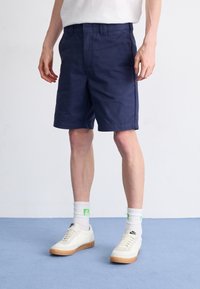 Pantaloni corti blu navy realizzati in misto cotone, con una vestibilità piatta e due tasche laterali, abbinati a sneakers bianche e calzini con accenti verdi.
