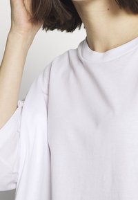 Light pink oversized t-shirt with a soft texture, wide sleeves, and a high neckline, partially shown in a close-up view.