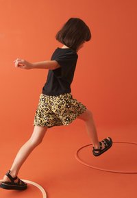Sort t-shirt kombineret med gule blomstrede shorts, der har en volangkant, og sorte kraftige sandaler, sat mod en orange baggrund.