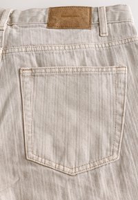 Jean en denim beige à motif chevrons, avec une étiquette de marque en cuir marron et une seule poche arrière ornée de coutures détaillées.