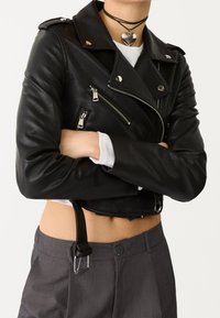Veste de moto en cuir noir, courte, avec des garnitures en argent, des poches zippées et un col pointu. Portée avec un pantalon gris et un haut blanc.