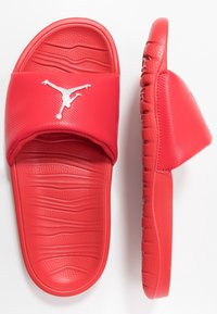 Punat punaiset sandaalit, joissa on uritettu hihna, kohokuvioitu logo ja muotoiltu pohja aaltoilevalla kuvioinnilla pitävää otetta varten.